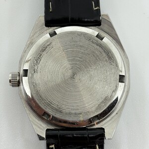 May include: A silver-toned watch case with a circular, brushed metal back featuring concentric grooves. The watch has a small, ridged winding crown on the left side and is attached to a black, textured leather strap with visible white stitching.