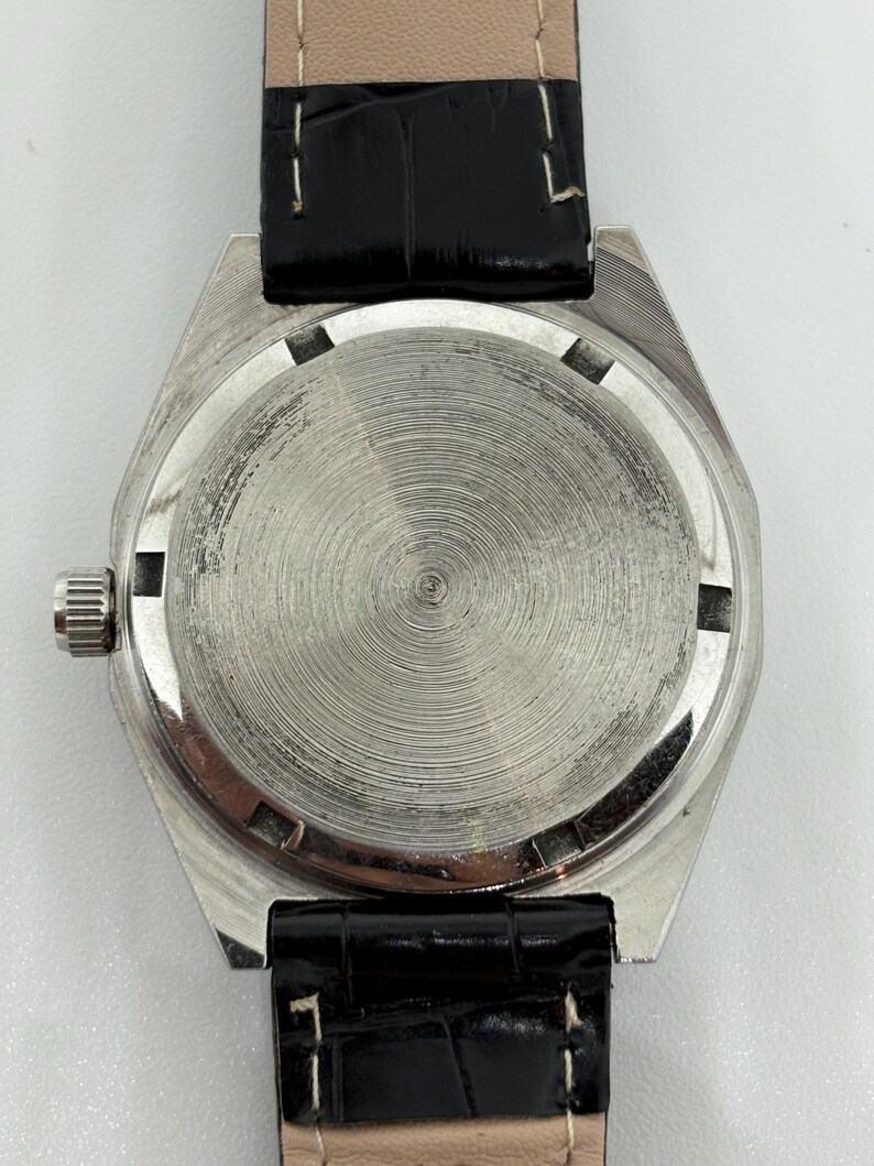 May include: A silver-toned watch case with a circular, brushed metal back featuring concentric grooves. The watch has a small, ridged winding crown on the left side and is attached to a black, textured leather strap with visible white stitching.