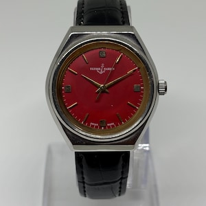 May include: A wristwatch with a silver-tone case, a vibrant red dial, and a black leather strap. The dial features gold-tone hour markers, gold-tone hands, and the Ulysse Nardin logo with an anchor symbol. The text Swiss Made is printed at the bottom.