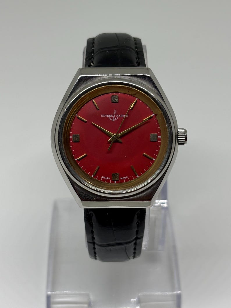 May include: A wristwatch with a silver-tone case, a vibrant red dial, and a black leather strap. The dial features gold-tone hour markers, gold-tone hands, and the Ulysse Nardin logo with an anchor symbol. The text Swiss Made is printed at the bottom.