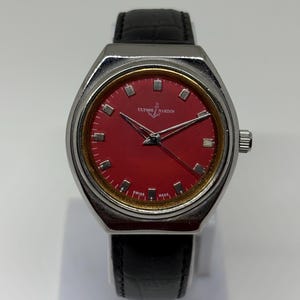 May include: A Ulysse Nardin wristwatch featuring a vibrant red dial with silver-tone hour markers, silver hands, and a gold-tone inner bezel. The watch has a polished silver-tone case and a black leather strap with a textured finish.