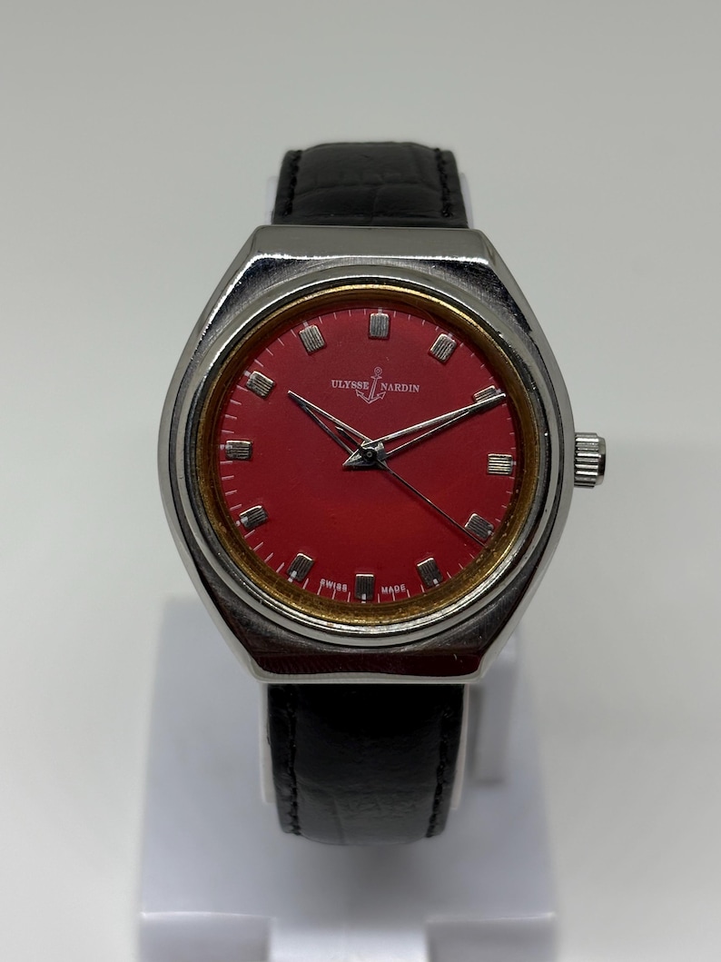 May include: A Ulysse Nardin wristwatch featuring a vibrant red dial with silver-tone hour markers, silver hands, and a gold-tone inner bezel. The watch has a polished silver-tone case and a black leather strap with a textured finish.