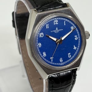 May include: An Ulysse Nardin wristwatch with a vibrant blue dial, white Arabic numerals, and silver-toned hands. The watch features a silver-toned metal case and a black textured leather strap with a crocodile-style pattern and a metal buckle.