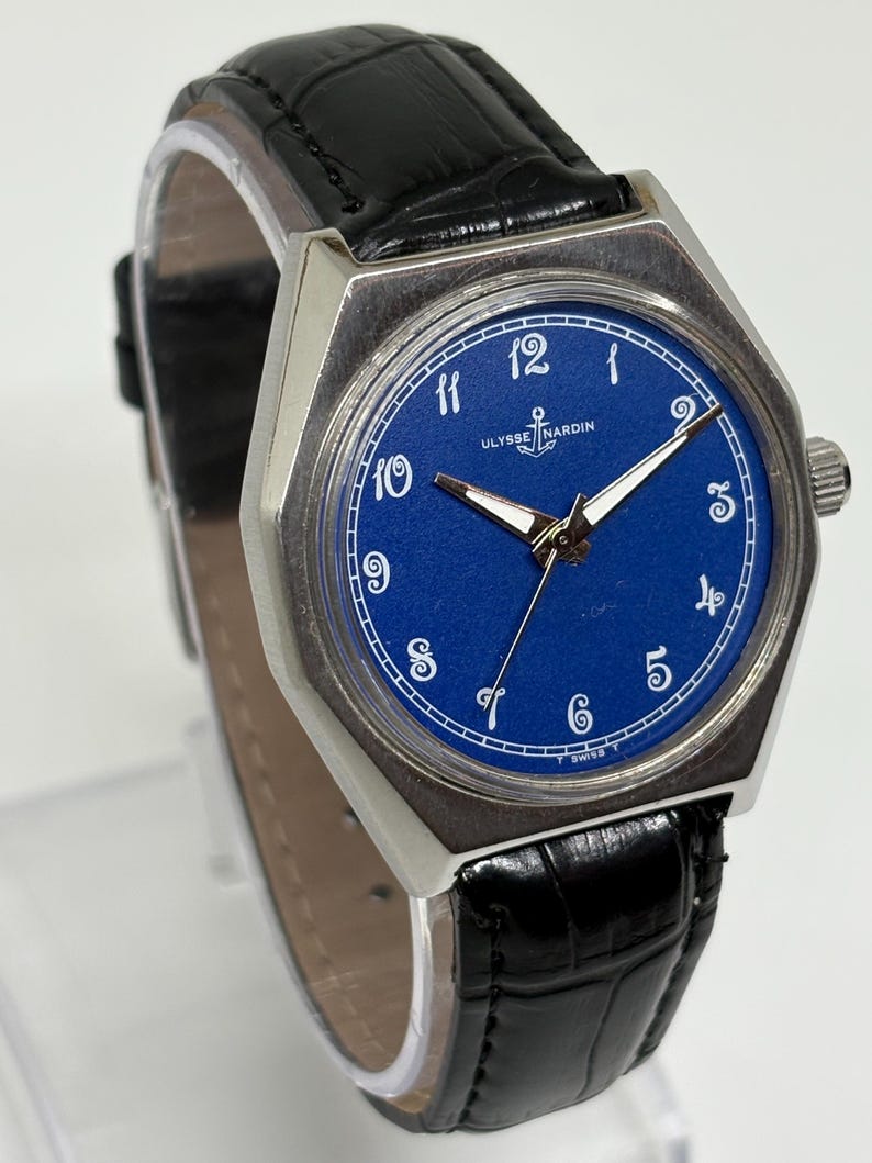 May include: An Ulysse Nardin wristwatch with a vibrant blue dial, white Arabic numerals, and silver-toned hands. The watch features a silver-toned metal case and a black textured leather strap with a crocodile-style pattern and a metal buckle.