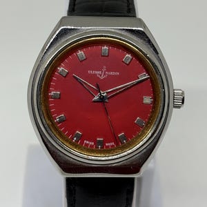 May include: A wristwatch with a silver-toned case, a vibrant red dial, and a black leather strap. The dial features silver hour markers, silver hands, and the text Ulysse Nardin with an anchor logo above the centre and Swiss Made at the bottom.