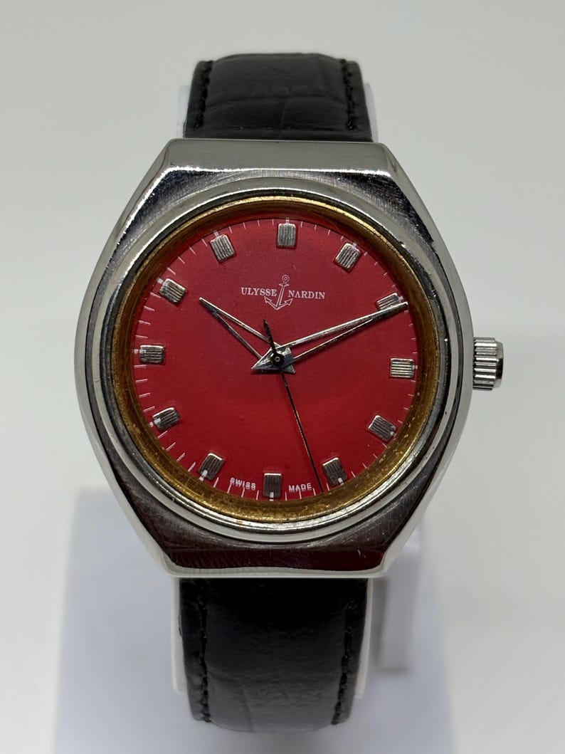 May include: A wristwatch with a silver-toned case, a vibrant red dial, and a black leather strap. The dial features silver hour markers, silver hands, and the text Ulysse Nardin with an anchor logo above the centre and Swiss Made at the bottom.