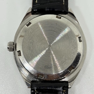 May include: A wristwatch with a circular silver-toned metal case back featuring a radial brushed finish and a notched perimeter. The watch is attached to a black leather strap with visible white stitching and a smooth, glossy surface texture.
