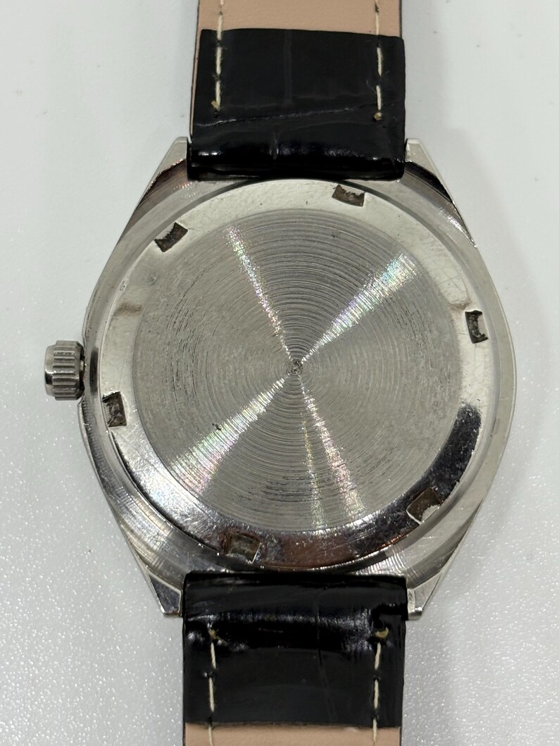 May include: A wristwatch with a circular silver-toned metal case back featuring a radial brushed finish and a notched perimeter. The watch is attached to a black leather strap with visible white stitching and a smooth, glossy surface texture.