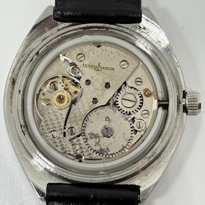 May include: A silver-toned wristwatch with an exposed mechanical movement, featuring a black leather strap. The movement is engraved with Ulysse Nardin, Swiss, and Seventeen Jewels. The internal gears, balance wheel, and mainspring are visible.