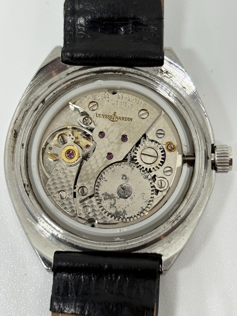 May include: A silver-toned wristwatch with an exposed mechanical movement, featuring a black leather strap. The movement is engraved with Ulysse Nardin, Swiss, and Seventeen Jewels. The internal gears, balance wheel, and mainspring are visible.
