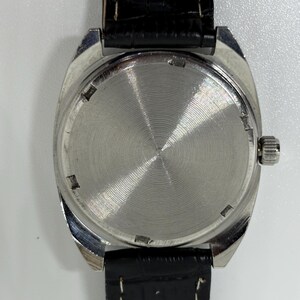 K&ouml;nnte beinhalten: Eine Armbanduhr mit einem runden, silberfarbenen Geh&auml;use mit geb&uuml;rsteter Metallr&uuml;ckseite und einem strukturierten schwarzen Lederarmband. Die Uhr verf&uuml;gt &uuml;ber eine kleine silberne Krone an der rechten Geh&auml;useseite und sichtbare wei&szlig;e N&auml;hte am Armband.