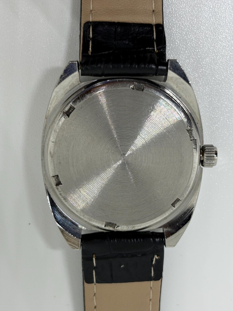K&ouml;nnte beinhalten: Eine Armbanduhr mit einem runden, silberfarbenen Geh&auml;use mit geb&uuml;rsteter Metallr&uuml;ckseite und einem strukturierten schwarzen Lederarmband. Die Uhr verf&uuml;gt &uuml;ber eine kleine silberne Krone an der rechten Geh&auml;useseite und sichtbare wei&szlig;e N&auml;hte am Armband.
