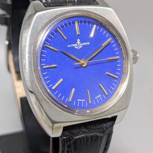May include: A vintage Ulysse Nardin watch with a square silver case and a vibrant blue dial. The watch features gold-colored hands and markers, and a black leather strap. The dial includes the text "Ulysse Nardin" and "Swiss Made".