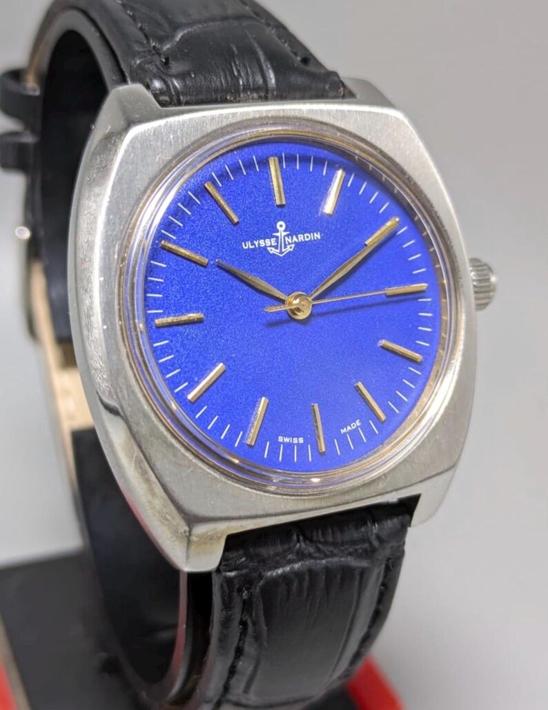 May include: A vintage Ulysse Nardin watch with a square silver case and a vibrant blue dial. The watch features gold-colored hands and markers, and a black leather strap. The dial includes the text "Ulysse Nardin" and "Swiss Made".