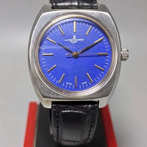 May include: A vintage Ulysse Nardin wristwatch with a silver case and a bright blue dial. The watch has gold hands and markers, a black leather strap, and a crown. It is displayed on a red and black stand. The watch is marked 'Swiss Made'.