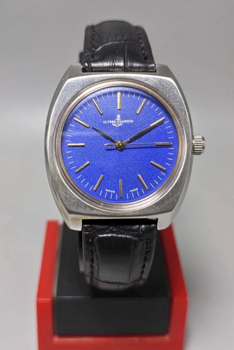 May include: A vintage Ulysse Nardin wristwatch with a silver-toned case and a vibrant blue dial. The watch features gold-colored hands and markers, a black leather strap, and a crown on the right side. The watch is displayed on a red and black stand.