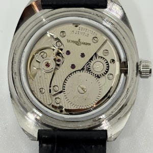 May include: A silver-toned wristwatch with the back casing removed to reveal the internal mechanical movement. The movement is engraved with Ulysse Nardin and 17 Jewels. The watch features a black leather strap and a metallic gear assembly.