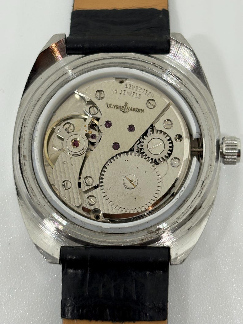 May include: A silver-toned wristwatch with the back casing removed to reveal the internal mechanical movement. The movement is engraved with Ulysse Nardin and 17 Jewels. The watch features a black leather strap and a metallic gear assembly.