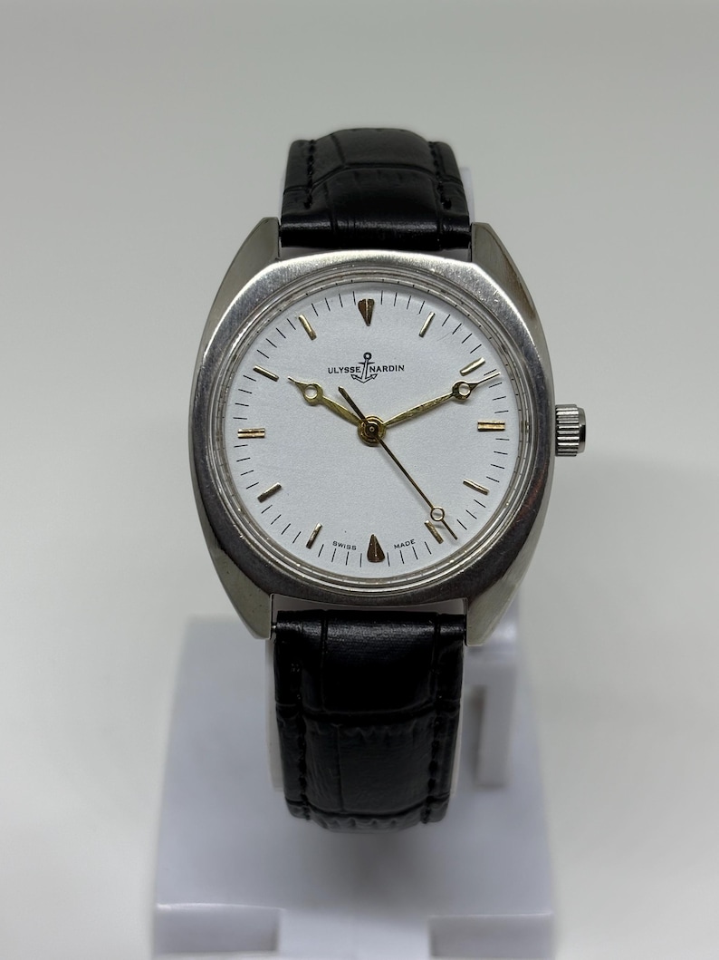 K&ouml;nnte beinhalten: Eine Ulysse Nardin Armbanduhr mit einem silberfarbenen Edelstahlgeh&auml;use, einem wei&szlig;en Zifferblatt sowie goldfarbenen Zeigern und Stundenmarkierungen. Die Uhr verf&uuml;gt &uuml;ber ein schwarzes Lederarmband mit Struktur und eine silberne Krone.