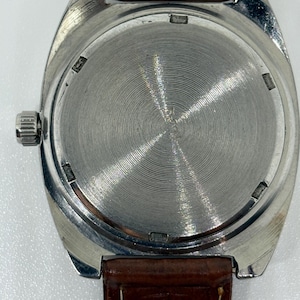 K&ouml;nnte beinhalten: Eine silberfarbene Armbanduhr mit rundem Geh&auml;use und einer geb&uuml;rsteten Metallr&uuml;ckseite mit konzentrischen kreisf&ouml;rmigen Rillen. Die Uhr ist an einem braunen Lederarmband mit sichtbaren N&auml;hten befestigt und hat eine Metallkrone an der Seite.