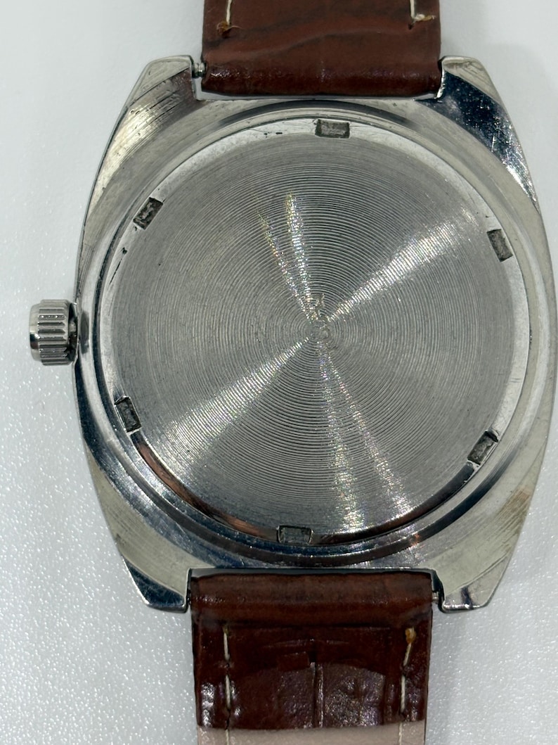 K&ouml;nnte beinhalten: Eine silberfarbene Armbanduhr mit rundem Geh&auml;use und einer geb&uuml;rsteten Metallr&uuml;ckseite mit konzentrischen kreisf&ouml;rmigen Rillen. Die Uhr ist an einem braunen Lederarmband mit sichtbaren N&auml;hten befestigt und hat eine Metallkrone an der Seite.