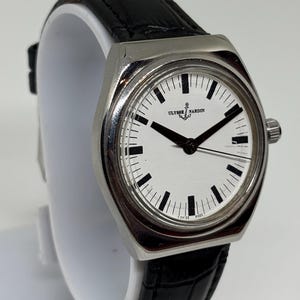 May include: A wristwatch with a silver-toned metal case and a white dial featuring black hour markers and hands. The dial displays the Ulysse Nardin logo with an anchor symbol. It is fitted with a black textured leather strap with a crocodile pattern.