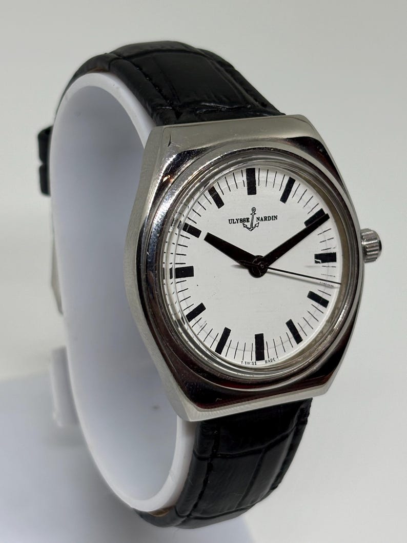 May include: A wristwatch with a silver-toned metal case and a white dial featuring black hour markers and hands. The dial displays the Ulysse Nardin logo with an anchor symbol. It is fitted with a black textured leather strap with a crocodile pattern.