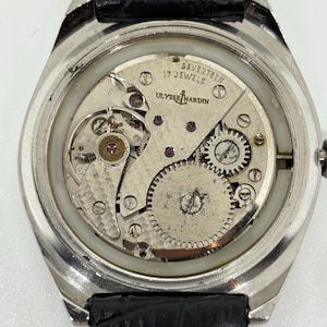 May include: A silver-toned wristwatch with the back casing removed to reveal the internal mechanical movement. The movement is engraved with ULYSSE NARDIN and SEVENTEEN 17 JEWELS. It features a black leather strap with visible white stitching.
