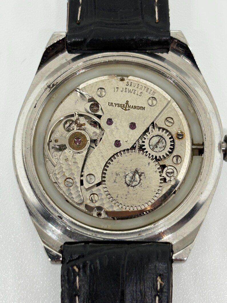 May include: A silver-toned wristwatch with the back casing removed to reveal the internal mechanical movement. The movement is engraved with ULYSSE NARDIN and SEVENTEEN 17 JEWELS. It features a black leather strap with visible white stitching.