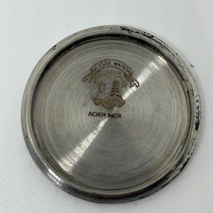 May include: A circular silver-toned metal watch case back with a brushed finish. The centre features an engraved Ulysse Nardin logo with a crest and banner, and the text ACIER INOX is stamped below the emblem on the flat metallic surface.