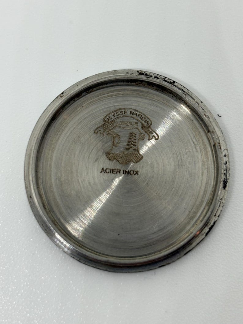 May include: A circular silver-toned metal watch case back with a brushed finish. The centre features an engraved Ulysse Nardin logo with a crest and banner, and the text ACIER INOX is stamped below the emblem on the flat metallic surface.