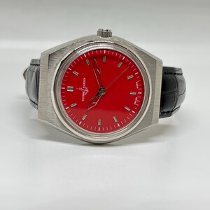 Puede incluir: Un reloj de pulsera Ulysse Nardin con esfera roja vibrante, marcadores de hora plateados y manecillas esqueletizadas. El reloj tiene una caja de acero inoxidable cepillado y una correa de cuero negro con textura. El texto Ulysse Nardin es visible.