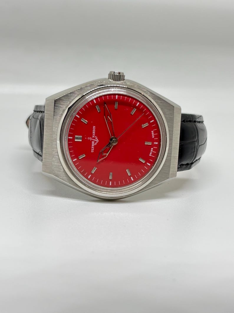 Puede incluir: Un reloj de pulsera Ulysse Nardin con esfera roja vibrante, marcadores de hora plateados y manecillas esqueletizadas. El reloj tiene una caja de acero inoxidable cepillado y una correa de cuero negro con textura. El texto Ulysse Nardin es visible.