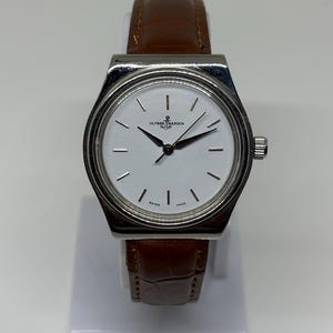 May include: A wristwatch with a silver-toned case and a white dial featuring thin black hour markers and hands. The dial displays the Ulysse Nardin logo with an anchor symbol and the text Swiss Made. It has a brown leather strap with a textured finish.