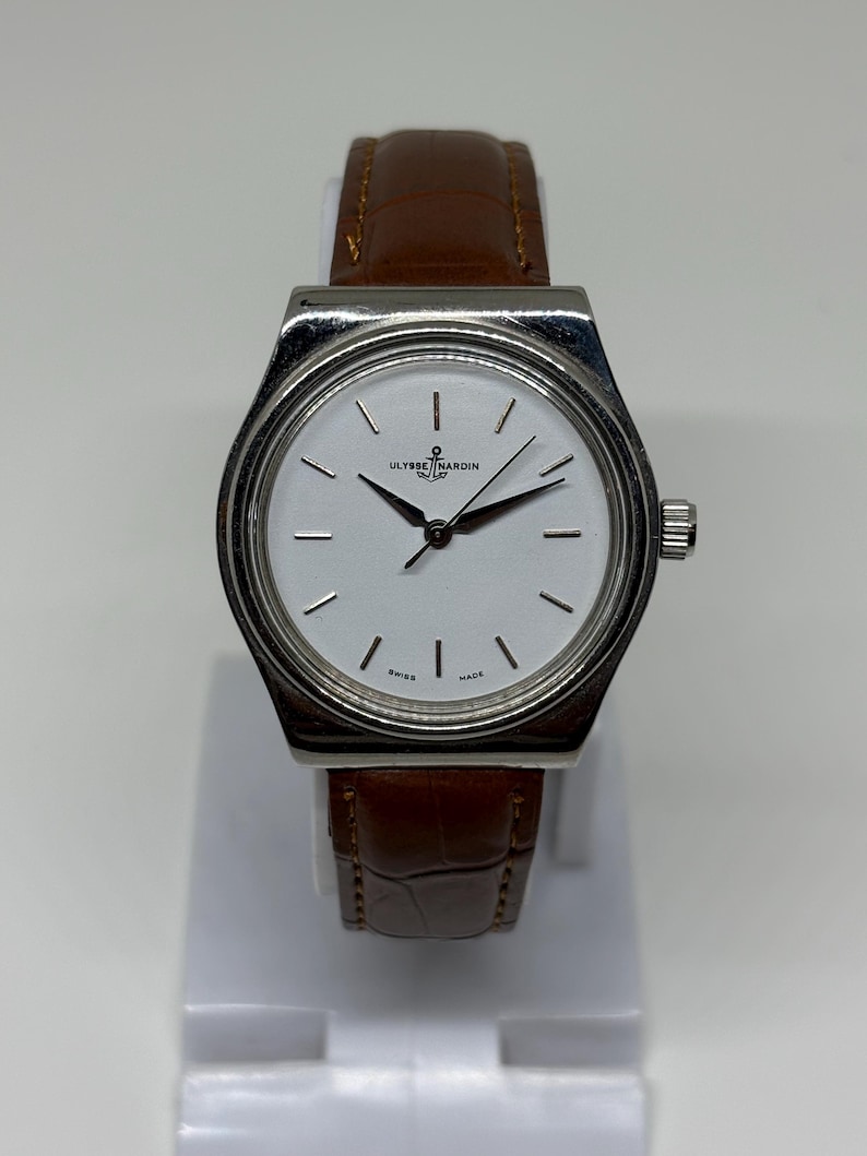 May include: A wristwatch with a silver-toned case and a white dial featuring thin black hour markers and hands. The dial displays the Ulysse Nardin logo with an anchor symbol and the text Swiss Made. It has a brown leather strap with a textured finish.