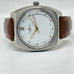 K&ouml;nnte beinhalten: Eine silberfarbene Ulysse Nardin Armbanduhr mit wei&szlig;em, strukturiertem Zifferblatt, goldfarbenen Zeigern, einer r&ouml;mischen Ziffer auf der Neun-Uhr-Position und einem braunen Lederarmband mit Krokodilpr&auml;gung und sichtbaren N&auml;hten.