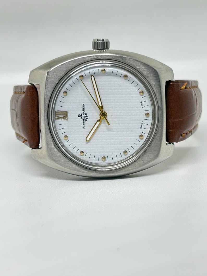 K&ouml;nnte beinhalten: Eine silberfarbene Ulysse Nardin Armbanduhr mit wei&szlig;em, strukturiertem Zifferblatt, goldfarbenen Zeigern, einer r&ouml;mischen Ziffer auf der Neun-Uhr-Position und einem braunen Lederarmband mit Krokodilpr&auml;gung und sichtbaren N&auml;hten.