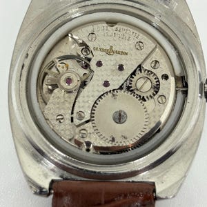 K&ouml;nnte beinhalten: Eine silberfarbene Uhr mit abgenommenem Geh&auml;useboden, die das interne mechanische Uhrwerk freilegt. Das Uhrwerk ist mit Ulysse Nardin, Swiss und 17 Jewels graviert. Ein braunes Lederarmband ist oben und unten am Uhrengeh&auml;use befestigt.
