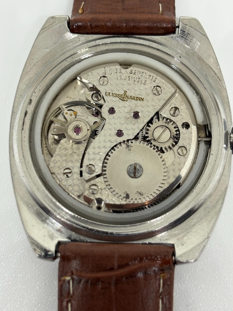 K&ouml;nnte beinhalten: Eine silberfarbene Uhr mit abgenommenem Geh&auml;useboden, die das interne mechanische Uhrwerk freilegt. Das Uhrwerk ist mit Ulysse Nardin, Swiss und 17 Jewels graviert. Ein braunes Lederarmband ist oben und unten am Uhrengeh&auml;use befestigt.
