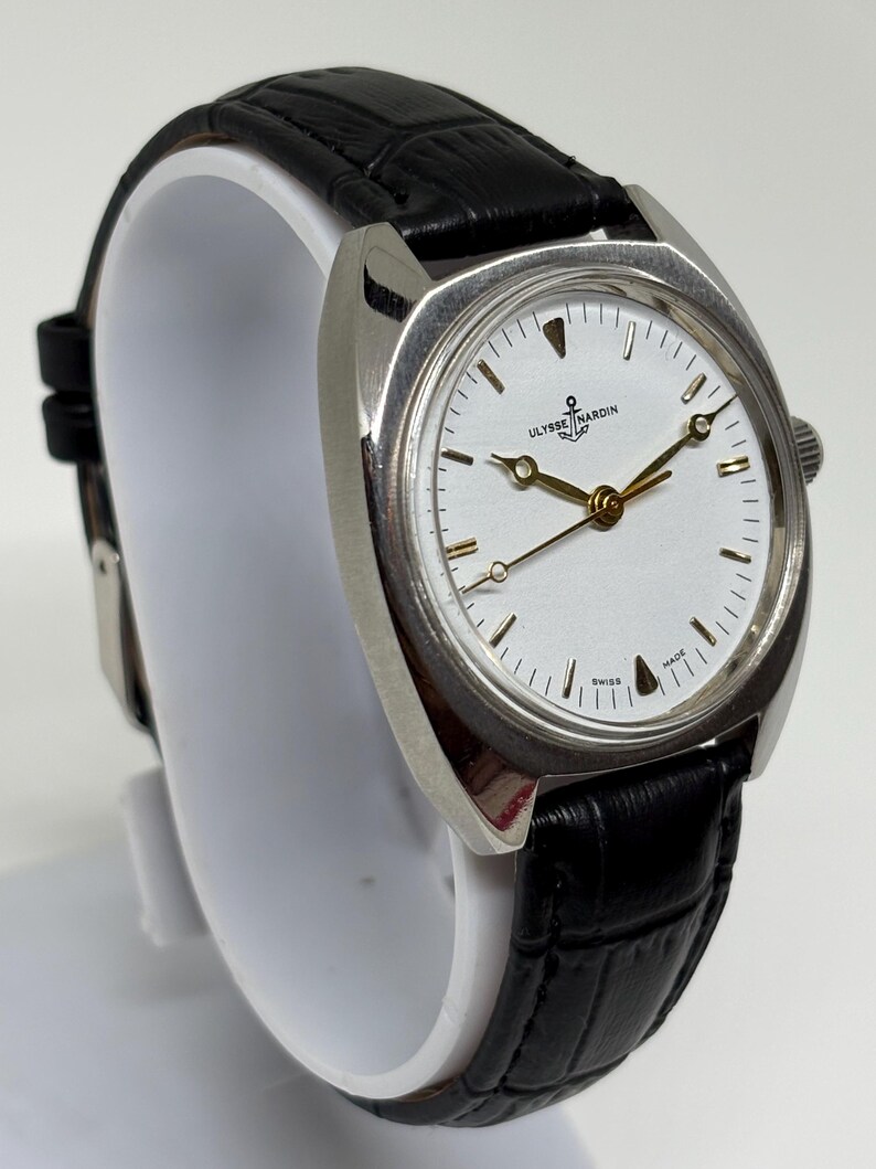 K&ouml;nnte beinhalten: Eine silberfarbene Armbanduhr mit wei&szlig;em Zifferblatt, goldfarbenen Zeigern und einem Anker-Logo mit dem Schriftzug ULYSSE NARDIN. Die Uhr hat ein schwarzes Lederarmband mit Strukturmuster und eine silberfarbene Krone an der Geh&auml;useseite.