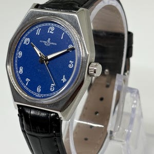 May include: A Ulysse Nardin wristwatch with a round blue dial, white Arabic numerals, and silver-tone hands. The watch features a silver-tone metal case and a black leather strap with a textured finish. The brand name and anchor logo are visible.