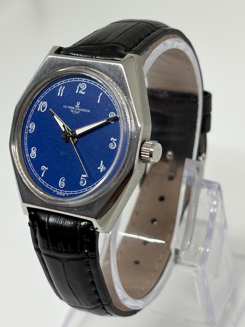 May include: A Ulysse Nardin wristwatch with a round blue dial, white Arabic numerals, and silver-tone hands. The watch features a silver-tone metal case and a black leather strap with a textured finish. The brand name and anchor logo are visible.