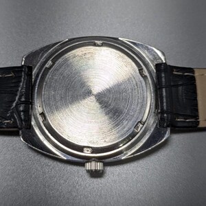 May include: Close-up of the back of a silver wristwatch with a black leather band. The watch face is circular with a textured, brushed metal finish. The band is black leather with a light tan underside.