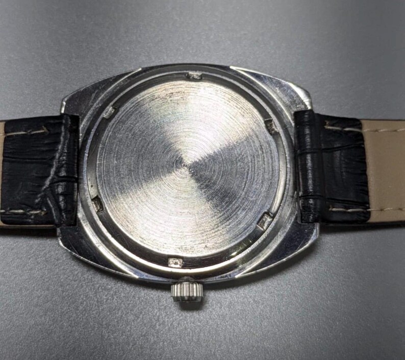 May include: Close-up of the back of a silver wristwatch with a black leather band. The watch face is circular with a textured, brushed metal finish. The band is black leather with a light tan underside.
