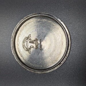 May include: A close-up of the back of a silver-colored watch. The watch back has a circular design with the words "ULYSSE NARDIN LOCLE" and "ACIER INOX" engraved on it. A crest is in the center.