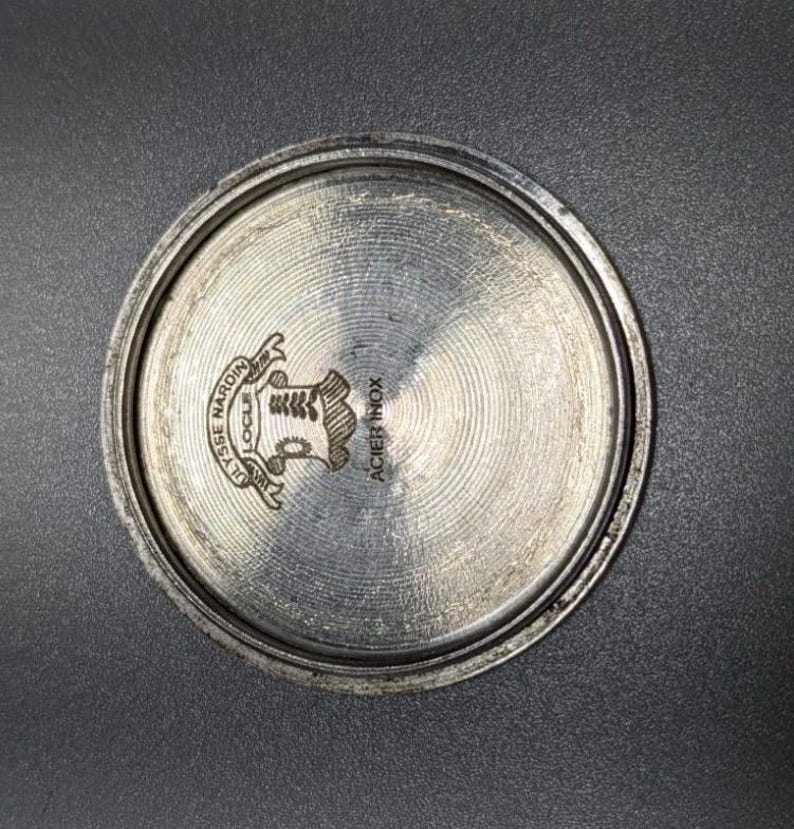 May include: A close-up of the back of a silver-colored watch. The watch back has a circular design with the words "ULYSSE NARDIN LOCLE" and "ACIER INOX" engraved on it. A crest is in the center.