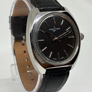May include: A Ulysse Nardin wristwatch featuring a round black dial with silver-tone hour markers and hands, housed in a polished silver-tone metal case. The watch is attached to a black textured leather strap with a visible buckle and pin holes.
