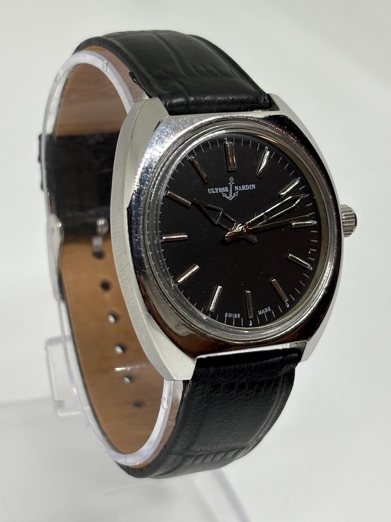 May include: A Ulysse Nardin wristwatch featuring a round black dial with silver-tone hour markers and hands, housed in a polished silver-tone metal case. The watch is attached to a black textured leather strap with a visible buckle and pin holes.