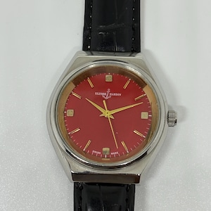 May include: A Ulysse Nardin wristwatch featuring a round silver-tone case, a vibrant red dial with gold-tone hour markers and hands, and a black leather strap with a textured finish. The dial displays the brand name and an anchor logo.