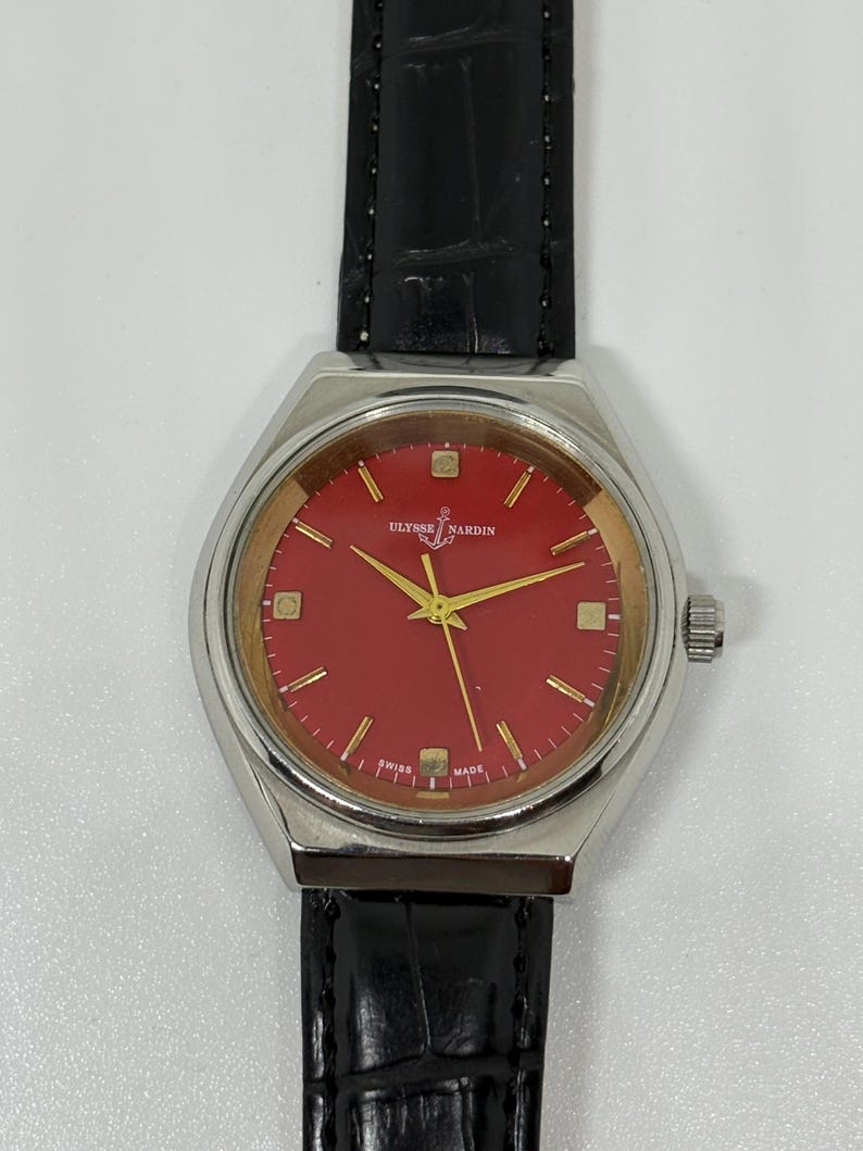 May include: A Ulysse Nardin wristwatch featuring a round silver-tone case, a vibrant red dial with gold-tone hour markers and hands, and a black leather strap with a textured finish. The dial displays the brand name and an anchor logo.
